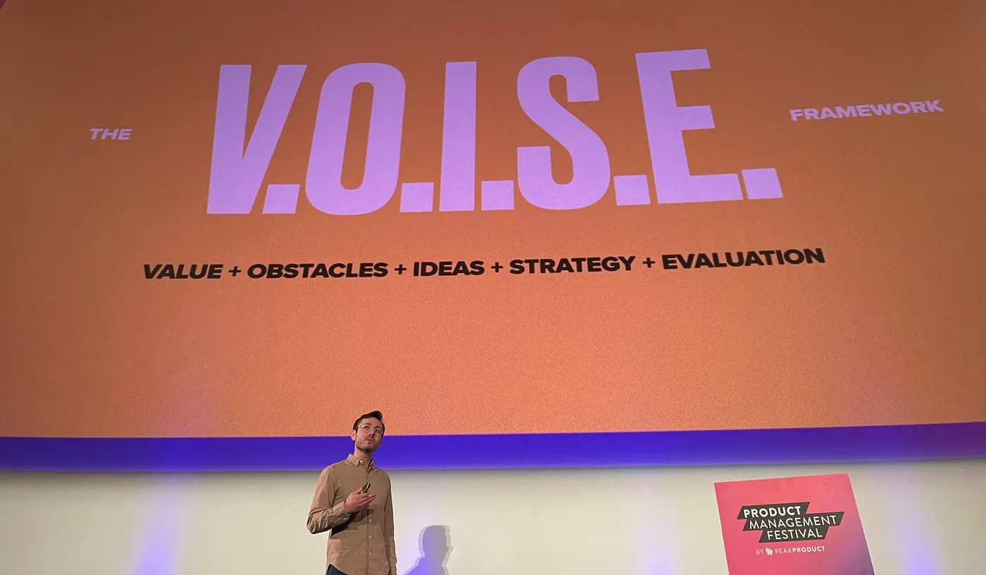 Ben presenting at Product Management Festival, in front of a slide reading “The V.O.I.S.E. framework: Value + Obstacles + Ideas + Strategy + Evaluation”.